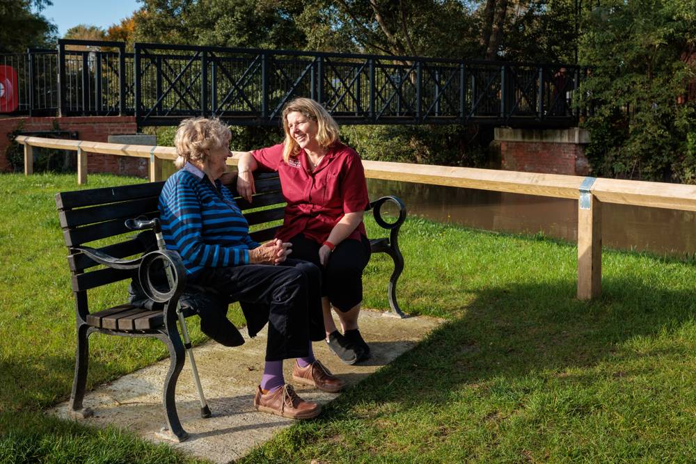 A carer and client laughing together on a park bench